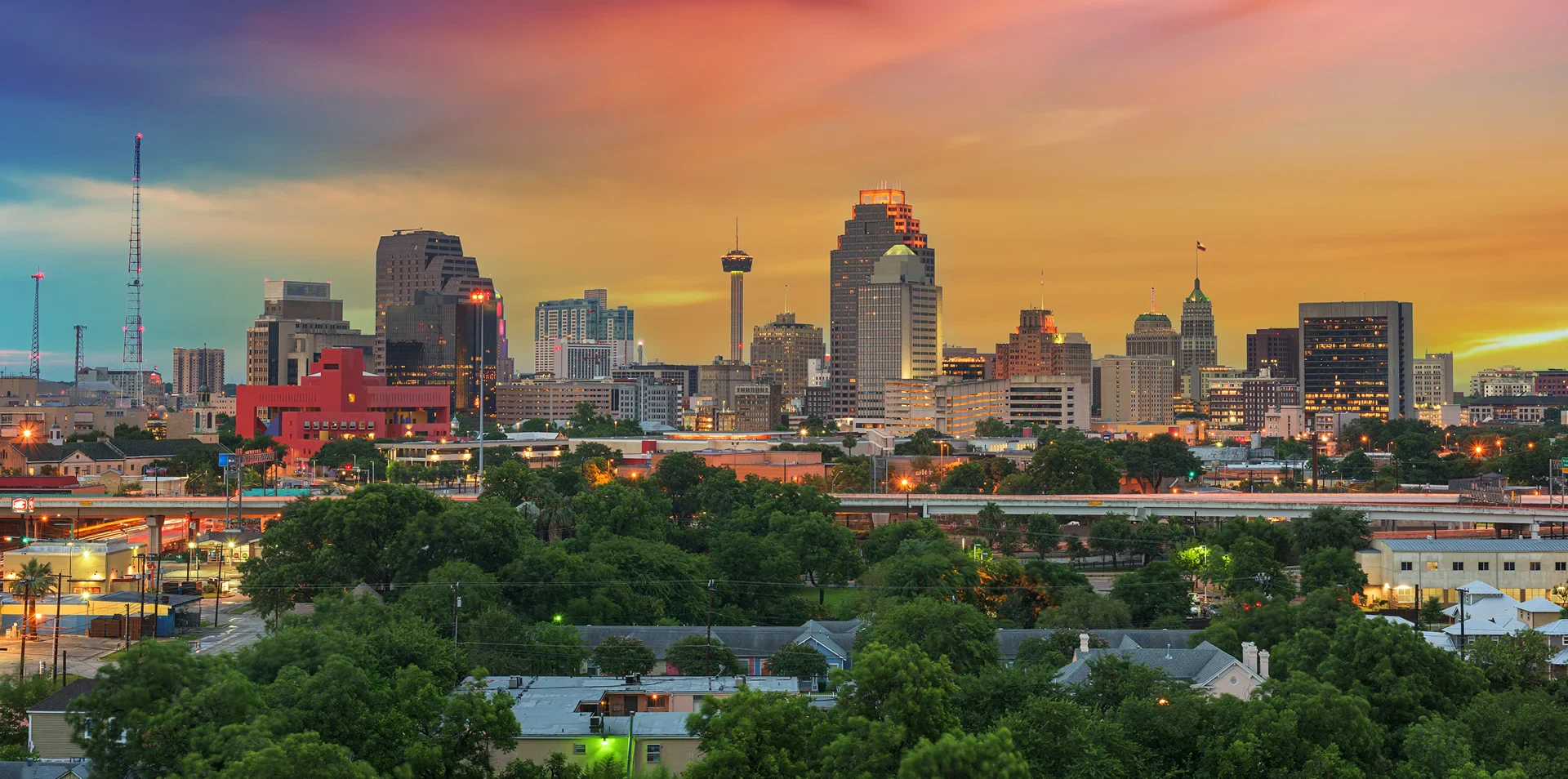 Sunset view of a city skyline with tall buildings, greenery in the foreground, and colorful sky hues