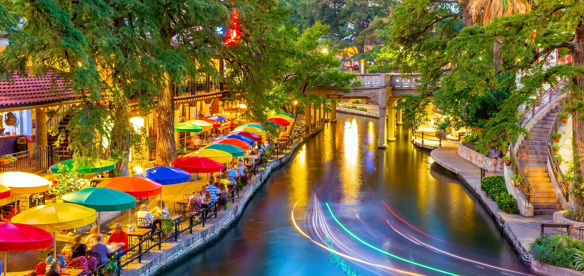 Evening view of a vibrant riverside walkway with colorful umbrellas, outdoor diners, lush trees, and a light-trail boat on the water