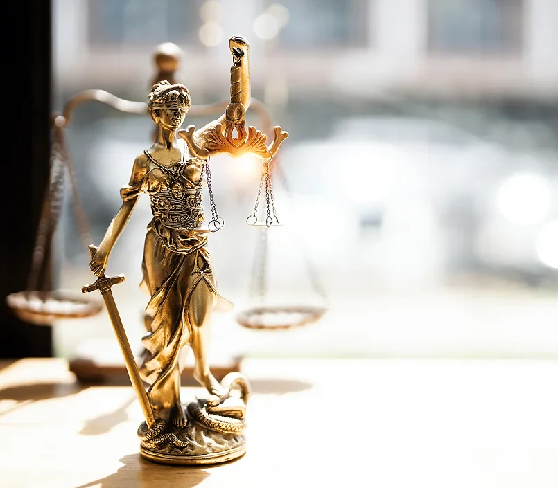 Golden Lady Justice statue holding scales and sword, with sunlight glowing through the scales, set against a blurred background